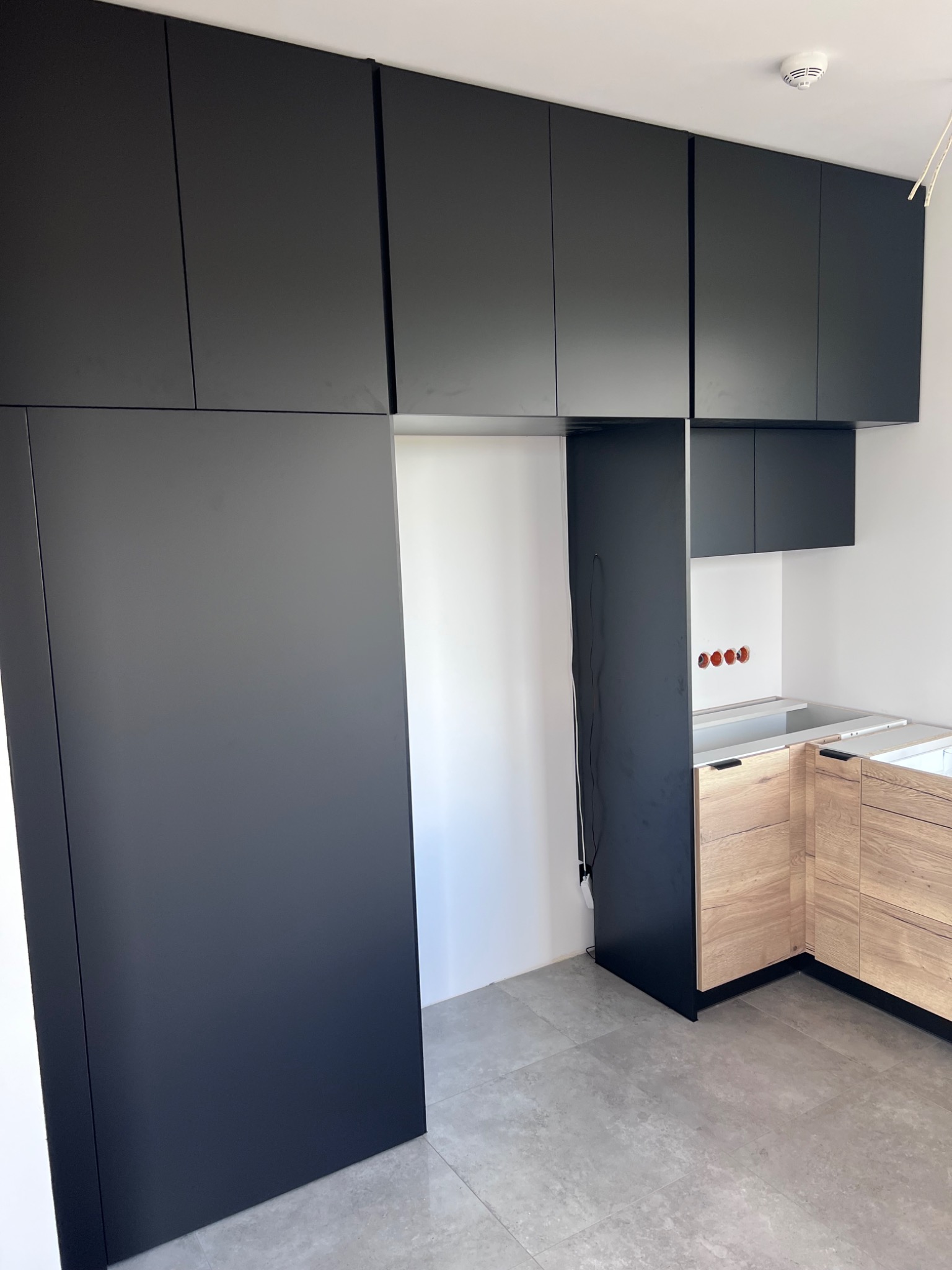 Częściowo zamontowana kuchnia na wymiar: czarne, matowe szafki wiszące i wysoka zabudowa obok drewnianych szafek dolnych z szufladami, widoczne gniazdka elektryczne w ścianie.