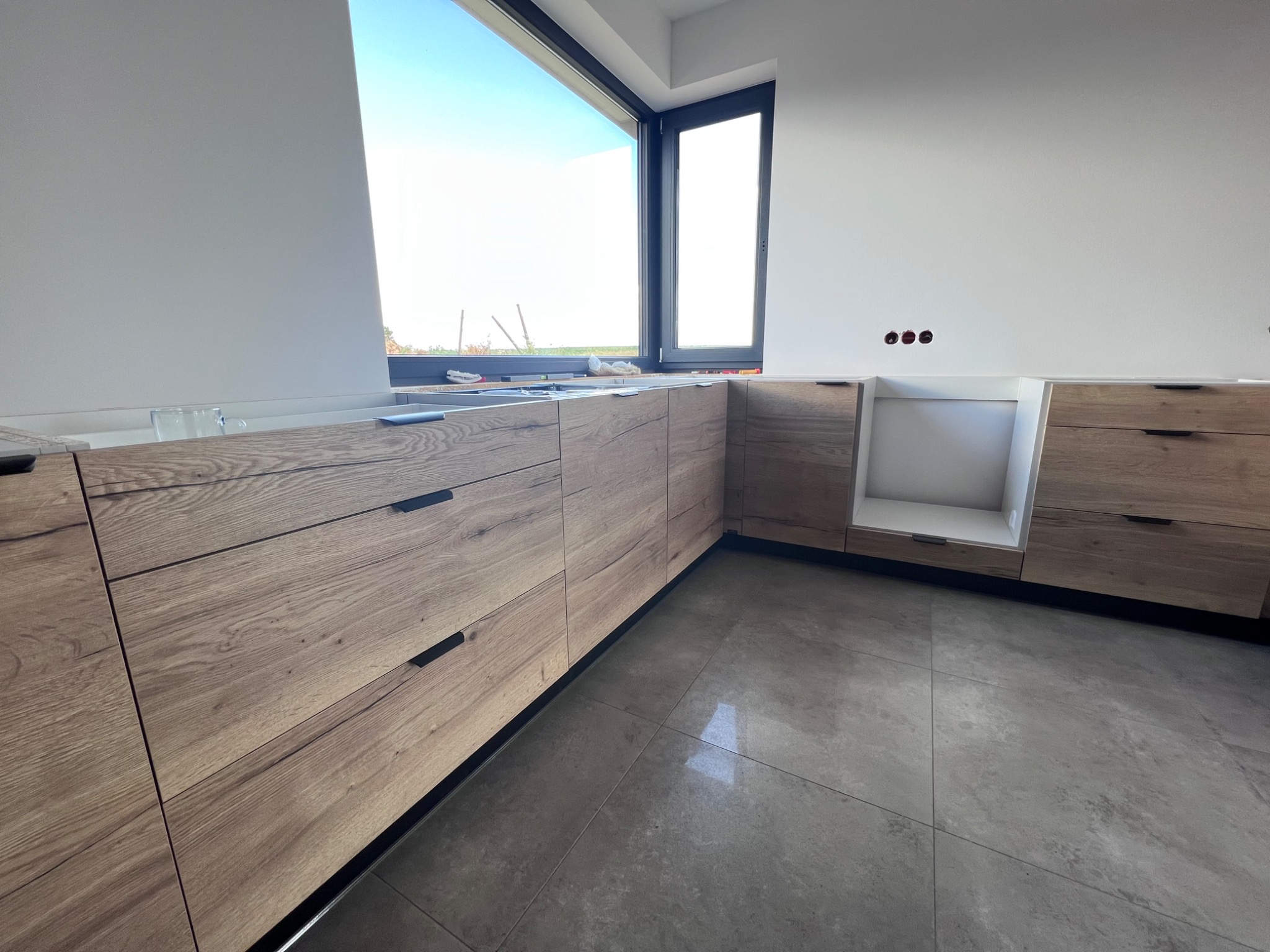 Fragment montażu kuchni: drewnopodobne fronty szuflad z czarnymi uchwytami, widoczne puszki elektryczne w ścianie, duże okno z widokiem na zewnątrz.