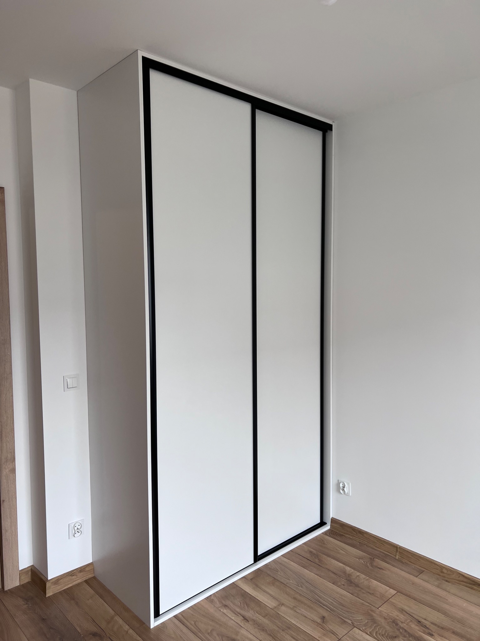 Biała szafa wnękowa z przesuwnymi drzwiami i czarnymi obramowaniami, dopasowana do wysokości pomieszczenia, z widocznym gniazdkiem elektrycznym i drewnianą podłogą