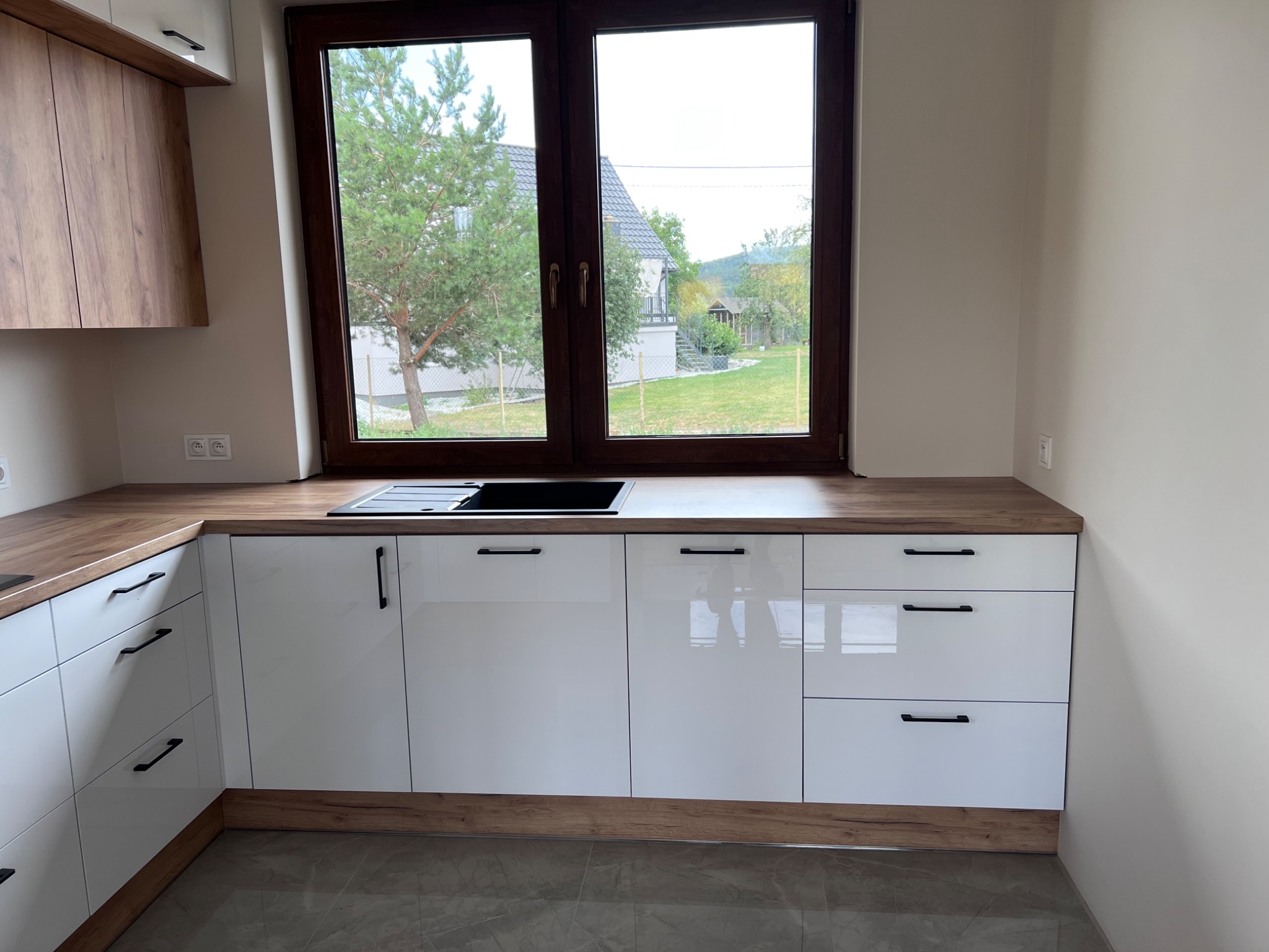 Biała, lakierowana zabudowa kuchenna z drewnianym blatem i frontami, czarne, poziome uchwyty, widok na ogród przez okno.