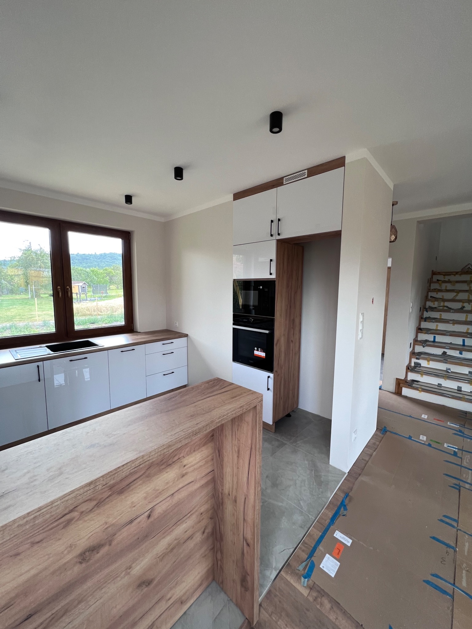 Wnętrze kuchni w trakcie montażu, widoczne białe szafki z czarnymi uchwytami, zabudowa z piekarnikiem i mikrofalówką w kolorze drewna, wyspa kuchenna z blatem drewnopodobnym, podłoga wyłożona...