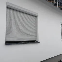 Zamknięta szara roleta zewnętrzna na oknie domu jednorodzinnego z widocznymi sopelkami lodu zwisającymi z dachu.