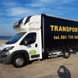 Biały bus dostawczy z czarną zabudową i napisem 'TRANSPORT' na tle plaży i morza, widoczne dodatkowe reflektory na dachu kabiny.