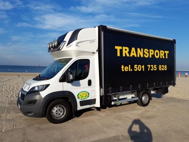 Biały bus dostawczy z czarną zabudową i napisem 'TRANSPORT' na tle plaży i morza, widoczne dodatkowe reflektory na dachu kabiny.