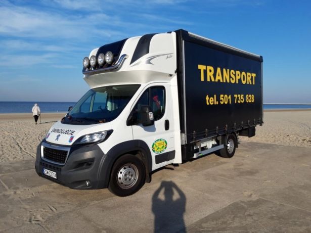 Biały bus z czarną plandeką i napisem 'TRANSPORT tel. 501 735 828' stoi na piaszczystej plaży, w tle morze i spacerująca osoba.