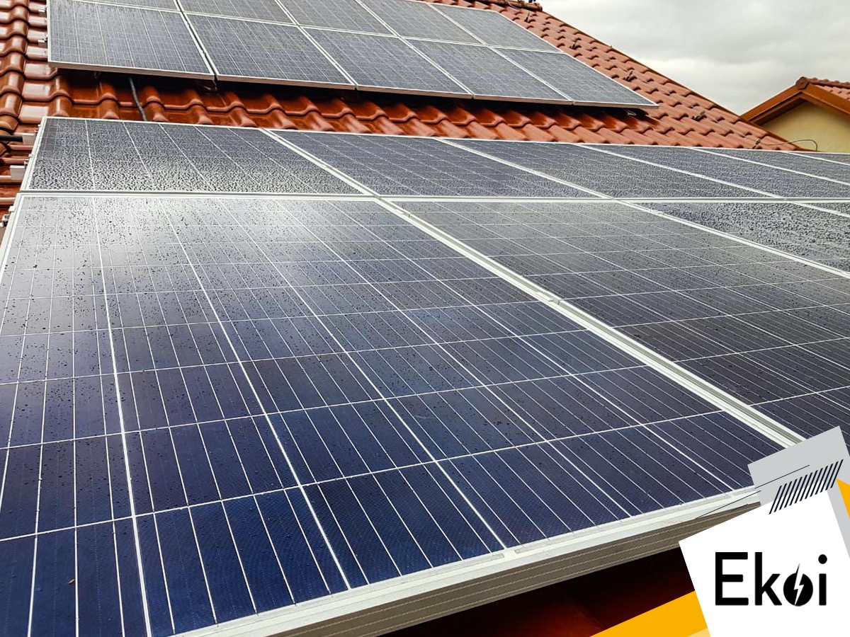 Panele słoneczne na czerwonym dachu pokryte kroplami deszczu, widoczne logo firmy Ekoi w prawym dolnym rogu.