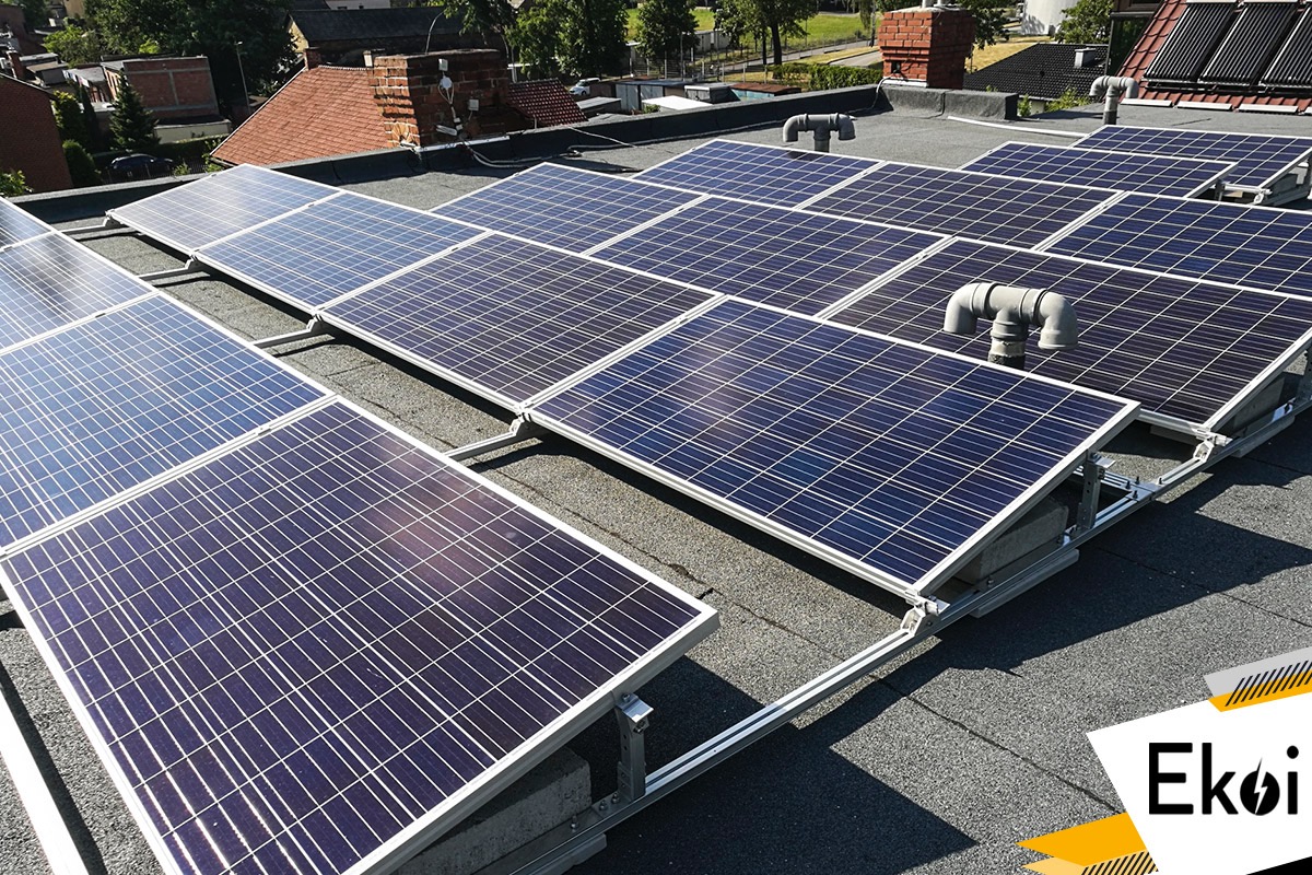Instalacja paneli słonecznych na płaskim dachu pokrytym papą, widoczne elementy mocujące i wentylacja, w tle budynki mieszkalne. W prawym dolnym rogu widoczne logo firmy Ekoi.
