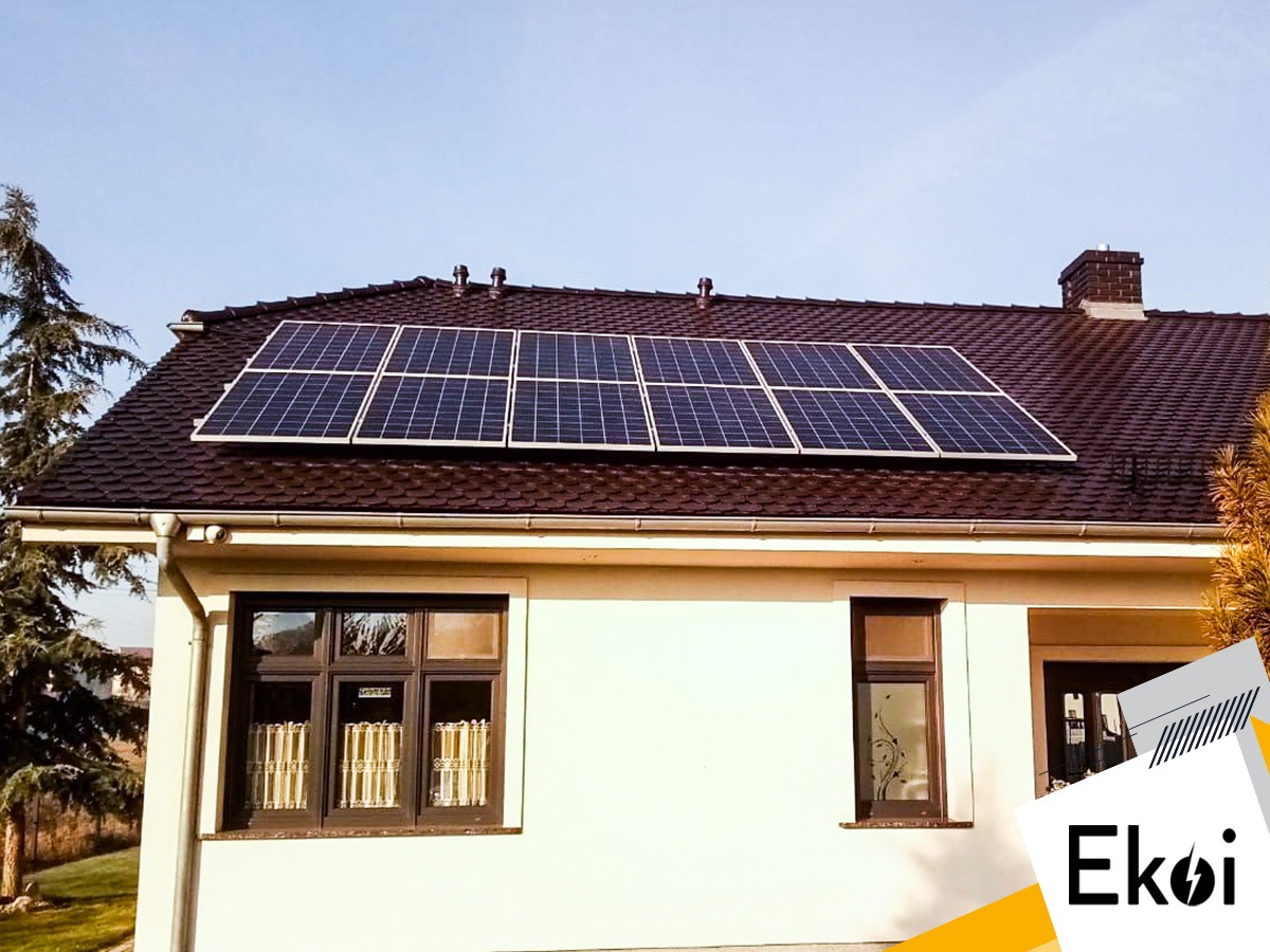 Panele fotowoltaiczne na dachu domu jednorodzinnego z brązową dachówką, widoczne okna i logo firmy Ekoi w prawym dolnym rogu.