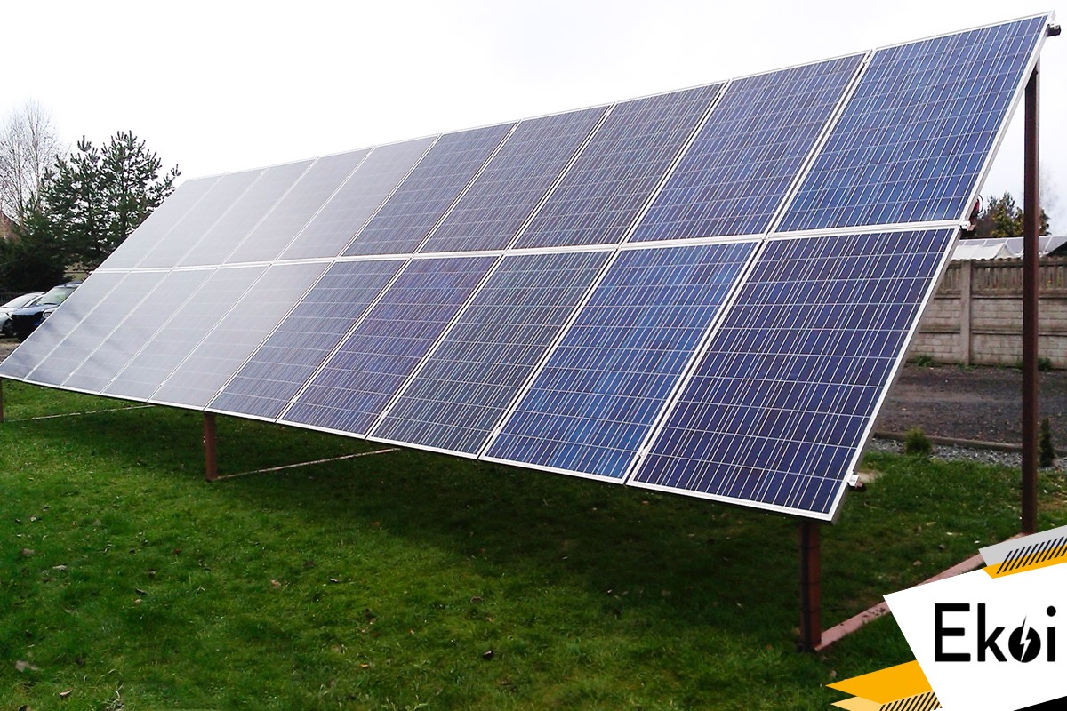 Instalacja paneli słonecznych na metalowych nogach, ustawiona na trawniku, z widocznym logo firmy Ekoi w prawym dolnym rogu.