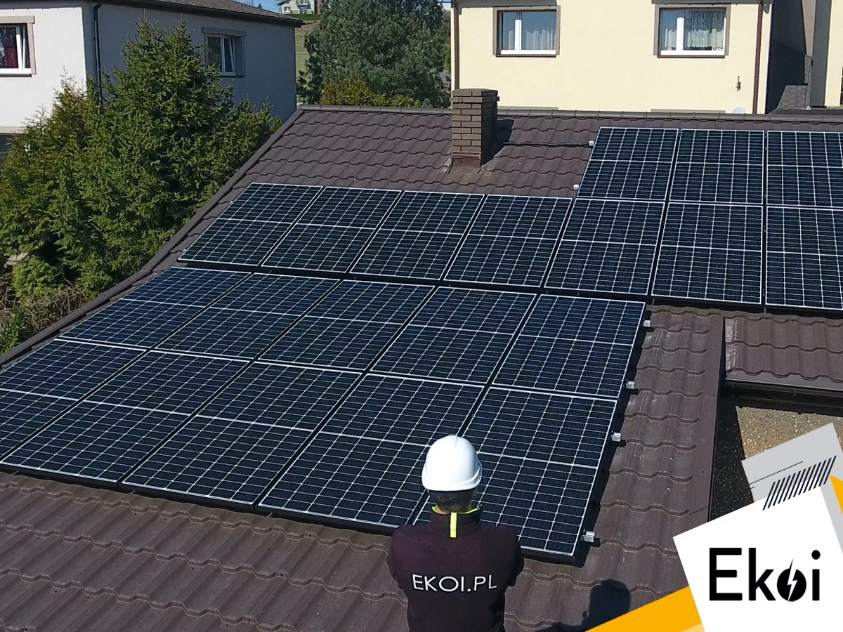 Montaż paneli słonecznych na brązowym dachu domu jednorodzinnego, widok z tyłu na pracownika w białym kasku i koszulce z logo firmy.