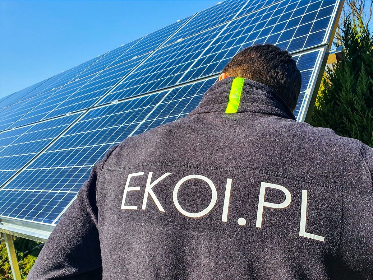 Instalator w polarze z logo EKOL.PL na tle paneli słonecznych pod błękitnym niebem.