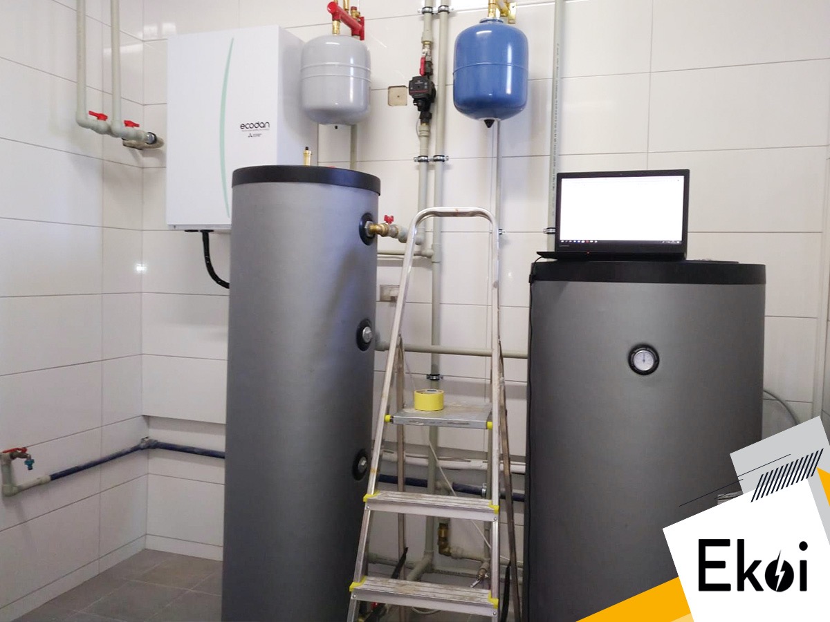 Instalacja z dwoma szarymi zbiornikami, białym urządzeniem Ecodan i niebieskim zbiornikiem wyrównawczym w pomieszczeniu wyłożonym białymi płytkami, z laptopem na jednym ze zbiorników i drabiną obok.