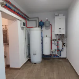 Instalacja grzewcza w kotłowni: dwa białe zbiorniki akumulacyjne, szary i niebieski zbiornik wyrównawczy, biały piec grzewczy na ścianie, rury w kolorach szarym, czerwonym i niebieskim...