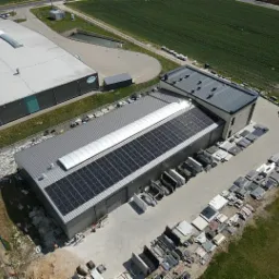 Widok z góry na budynek przemysłowy z panelami słonecznymi na dachu, obok składowane materiały budowlane, w tle pole uprawne i drugi budynek.