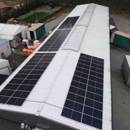 Przyjazna Energia.com. Sp.z o.o. - Widok z góry na biały dach hali z zamontowanymi panelami słonecznymi, obok zaparkowana ciężarówka z naczepą, w tle budynki i drzewa.