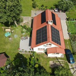 Przyjazna Energia.com. Sp.z o.o. - Widok z lotu ptaka na dom jednorodzinny z panelami słonecznymi na dachu, zadbany ogród z altaną i huśtawką, zaparkowany niebieski samochód.