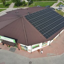 Przyjazna Energia.com. Sp.z o.o. - Widok z góry na budynek Delikatesów Centrum w Gdańsku z zainstalowanymi panelami słonecznymi na brązowym dachu wielospadowym. Na chodniku przed sklepem stoi osoba.