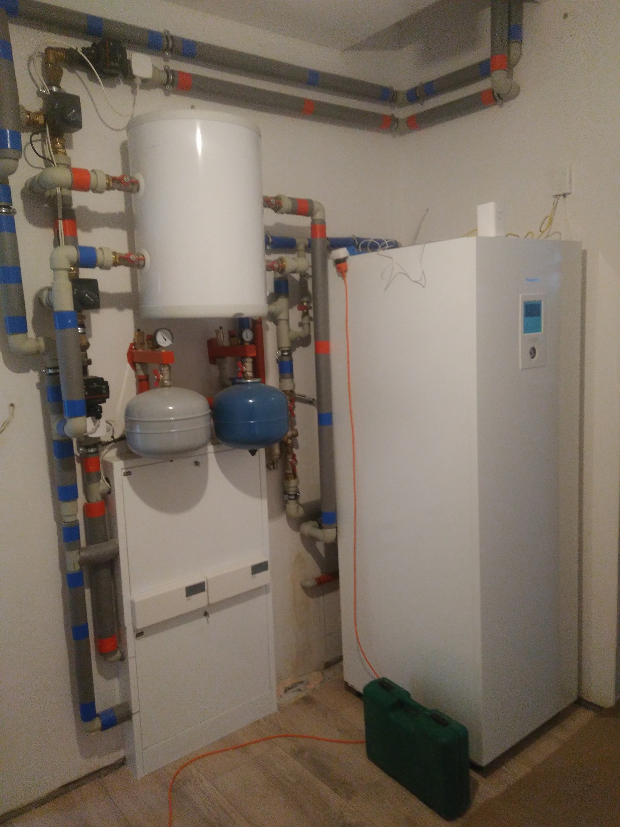 Kompletna instalacja centralnego ogrzewania z zbiornikiem buforowym, naczyniami wzbiorczymi i rozbudowaną siecią rur w kotłowni, z zieloną skrzynką narzędziową na podłodze.