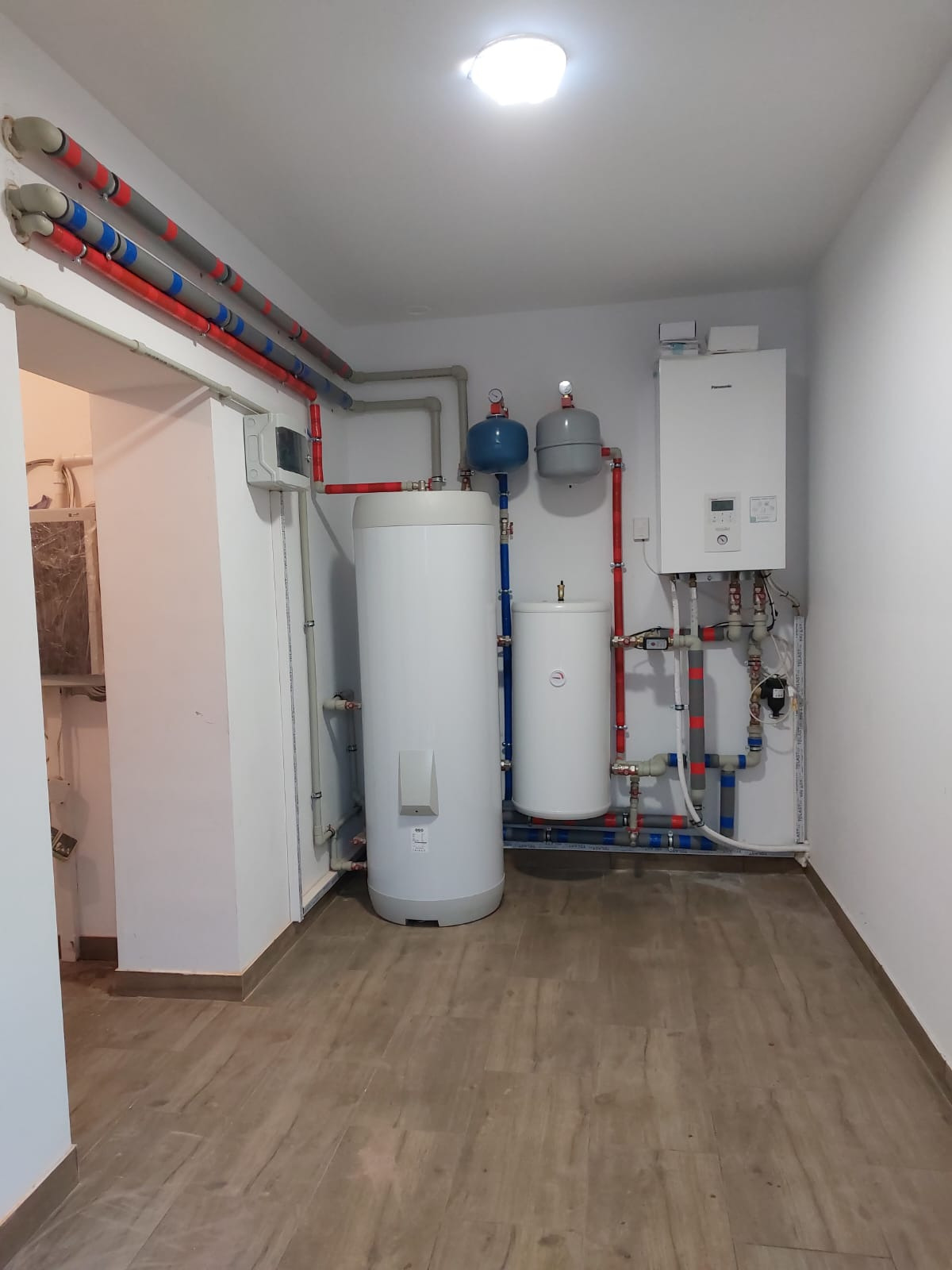 Instalacja grzewcza w kotłowni: dwa białe zbiorniki akumulacyjne, szary i niebieski zbiornik wyrównawczy, biały piec grzewczy na ścianie, rury w kolorach szarym, czerwonym i niebieskim...