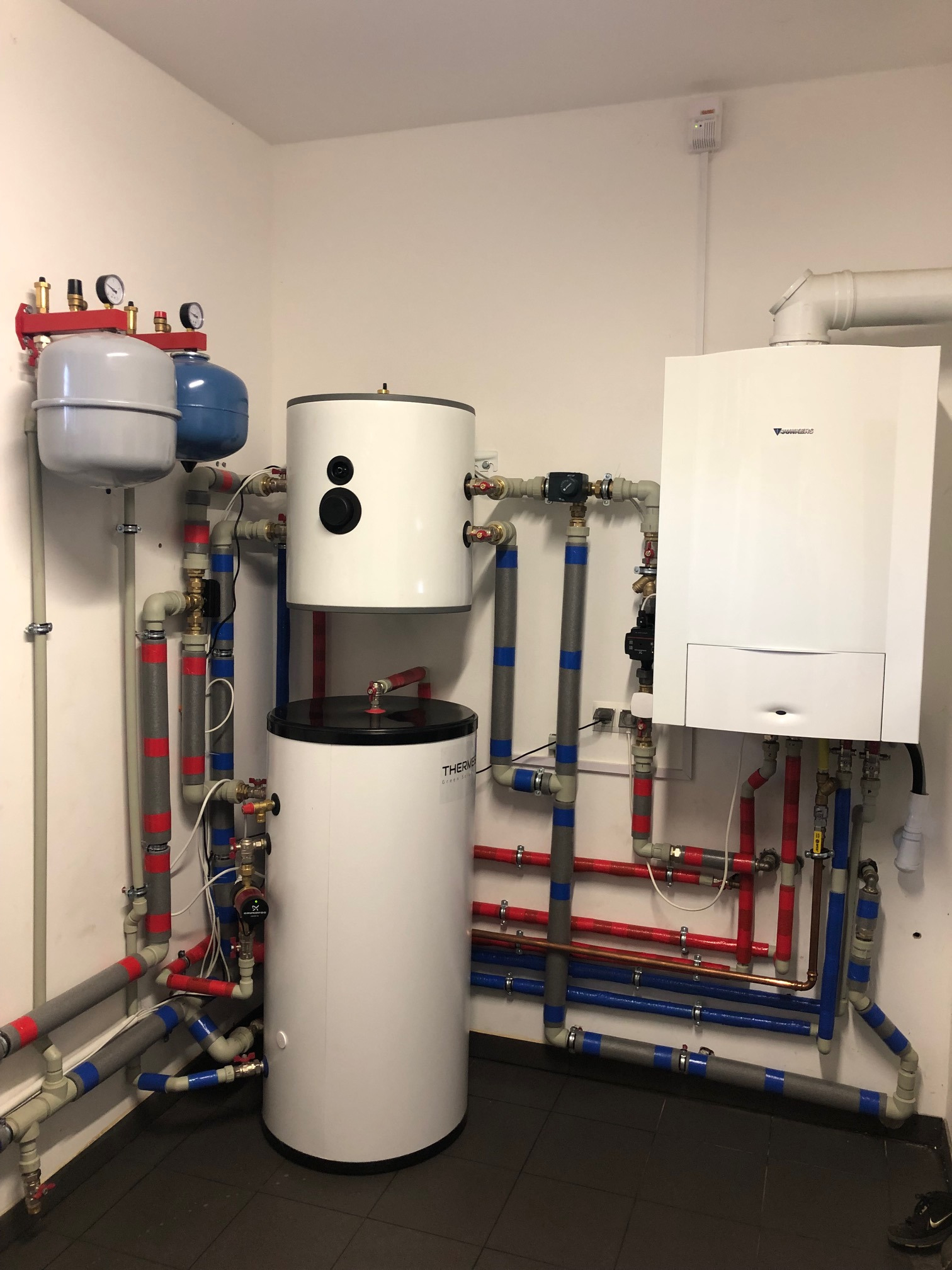 Kompletna instalacja kotłowni z zasobnikiem ciepłej wody, zbiornikami wyrównawczymi, pompą obiegową i kotłem gazowym Junkers, widoczne liczne rury w izolacji z oznaczeniami kolorystycznymi.