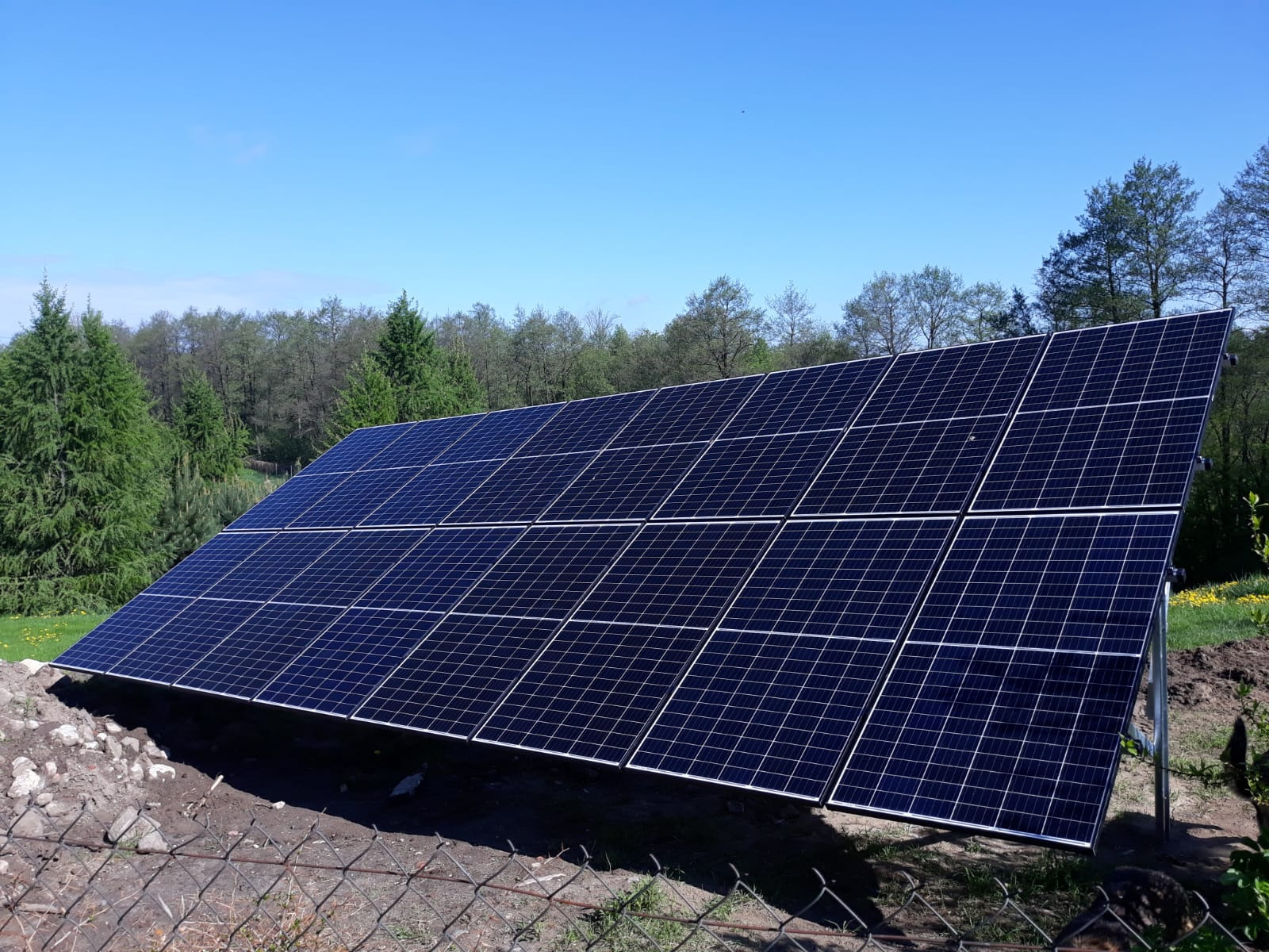 Panel fotowoltaiczny na metalowej konstrukcji, zamontowany na nierównym terenie z kamieniami, w tle widoczne drzewa i błękitne niebo.