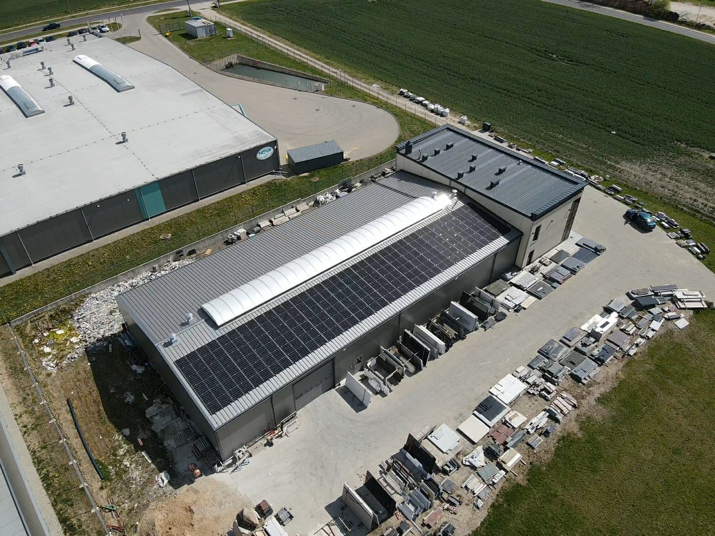 Widok z góry na budynek przemysłowy z panelami słonecznymi na dachu, obok składowane materiały budowlane, w tle pole uprawne i drugi budynek.