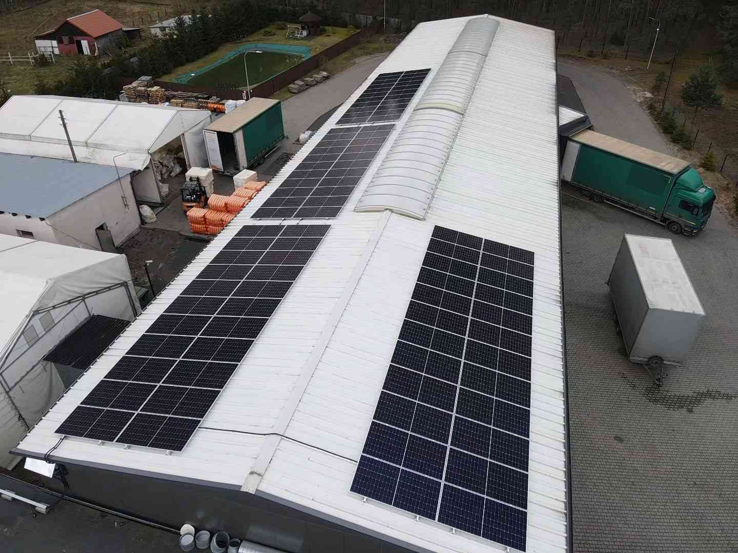 Widok z góry na biały dach hali z zamontowanymi panelami słonecznymi, obok zaparkowana ciężarówka z naczepą, w tle budynki i drzewa.