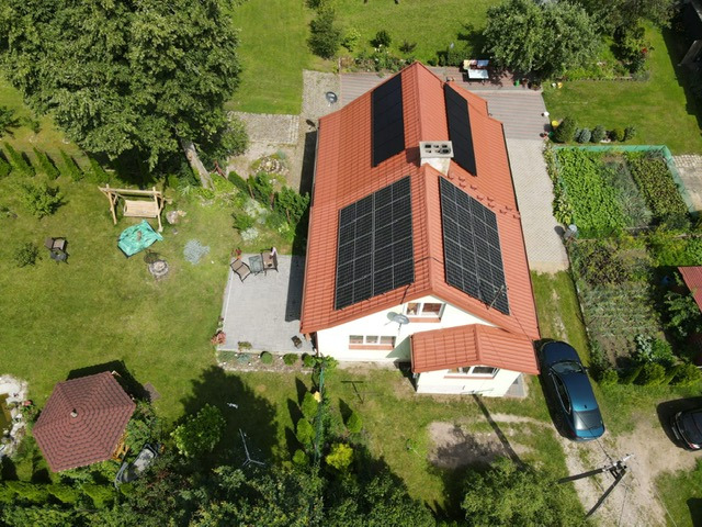 Widok z lotu ptaka na dom jednorodzinny z panelami słonecznymi na dachu, zadbany ogród z altaną i huśtawką, zaparkowany niebieski samochód.