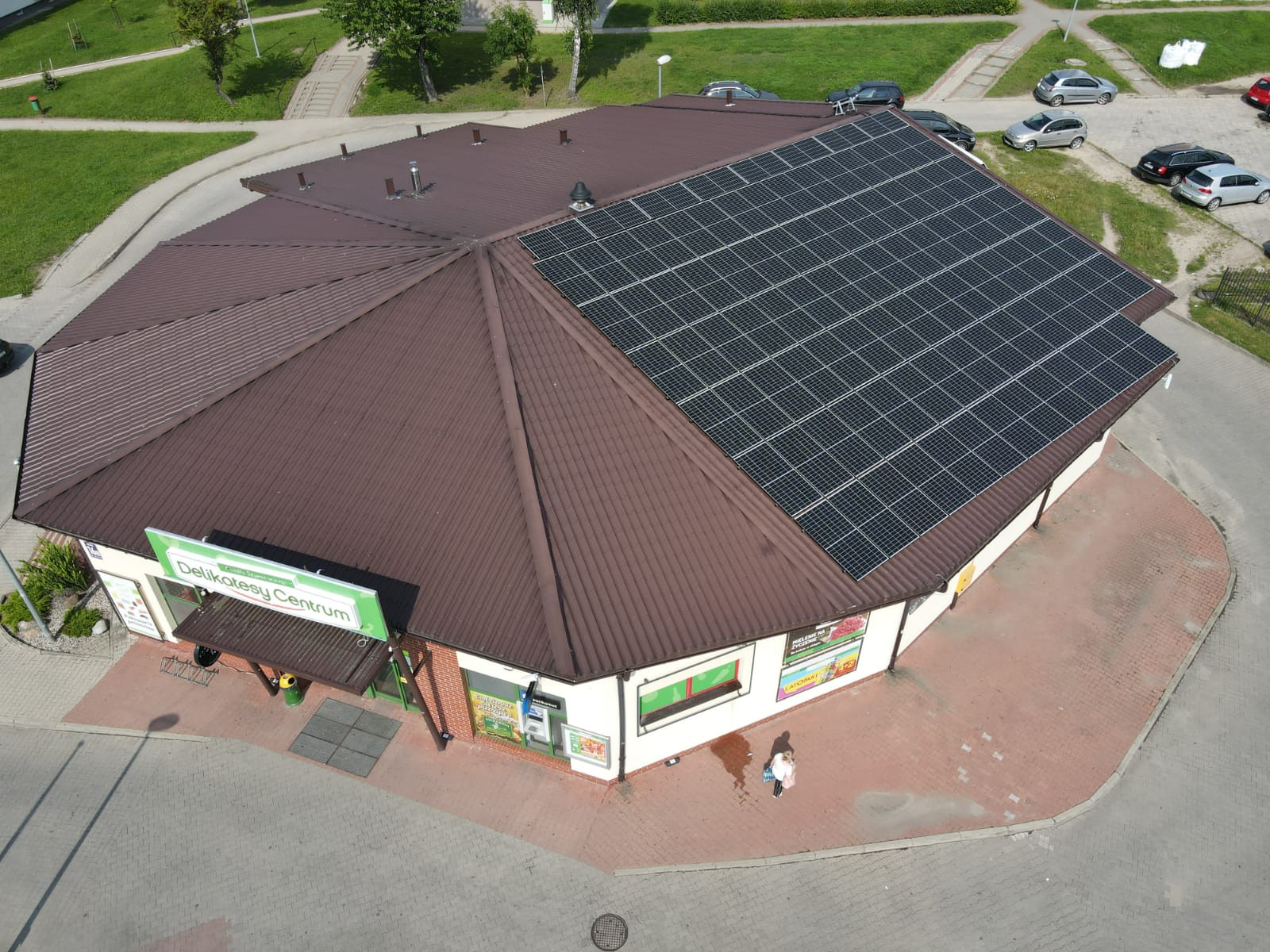 Widok z góry na budynek Delikatesów Centrum w Gdańsku z zainstalowanymi panelami słonecznymi na brązowym dachu wielospadowym. Na chodniku przed sklepem stoi osoba.