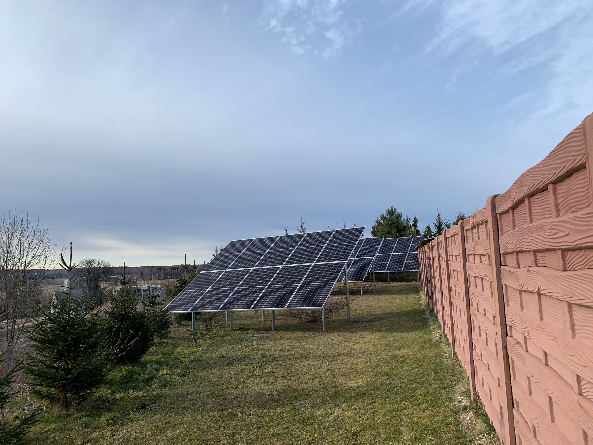 Rząd paneli słonecznych na trawiastym terenie, widoczny zza ozdobnego, betonowego ogrodzenia w kolorze terakoty, pod pochmurnym niebem.