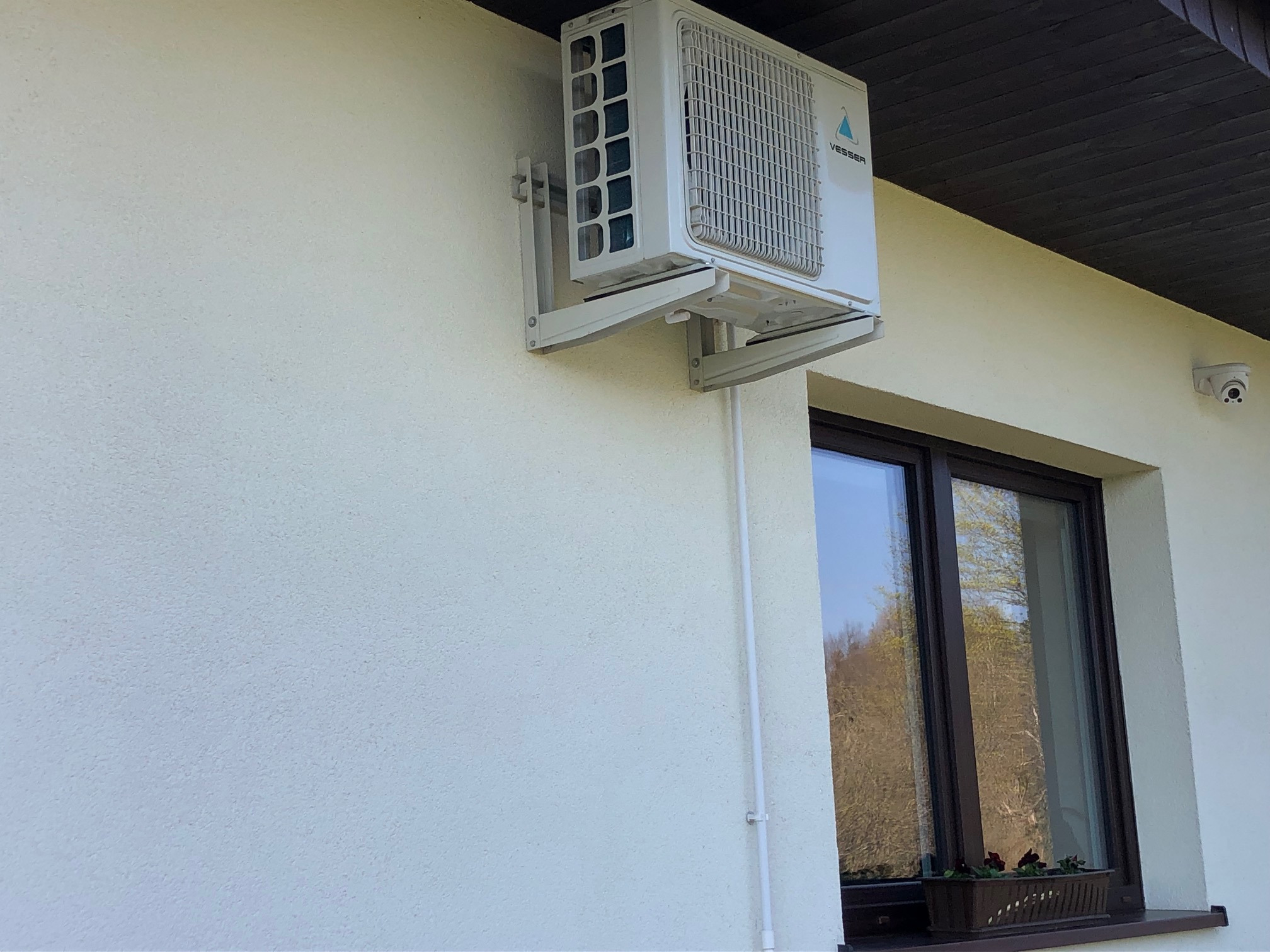 Biała jednostka zewnętrzna klimatyzatora marki Vesser zamontowana na ścianie budynku, widoczna kamera monitoringu, okno z doniczką z kwiatami.