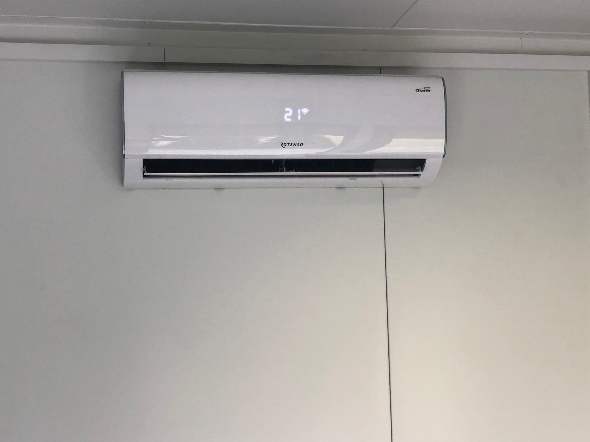 Biała klimatyzacja Rotenso z wyświetlaczem pokazującym 21 stopni Celsjusza zamontowana na jasnej ścianie.