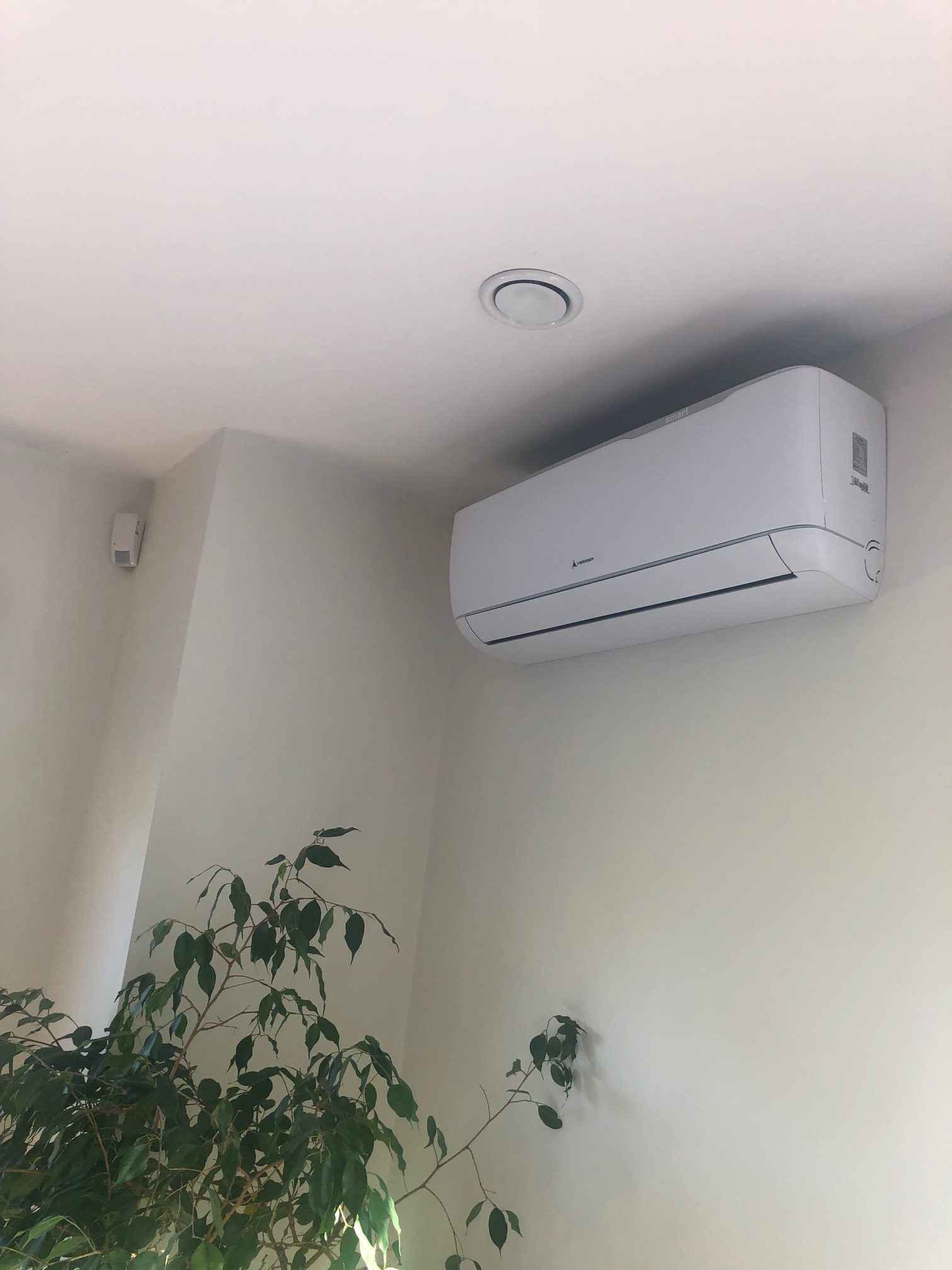 Biała jednostka klimatyzacji zamontowana na ścianie w jasnym pomieszczeniu, z widoczną rośliną doniczkową w rogu i okrągłym nawiewnikiem na suficie.