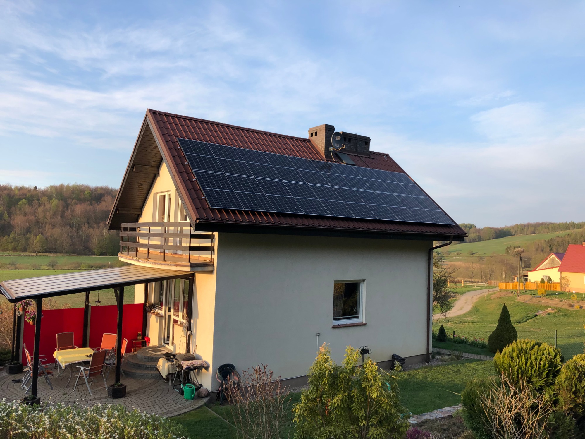 Dom jednorodzinny z panelami słonecznymi na dachu z czerwonej dachówki, widok z ogrodu w słoneczny dzień, zadaszony taras z meblami ogrodowymi z boku.