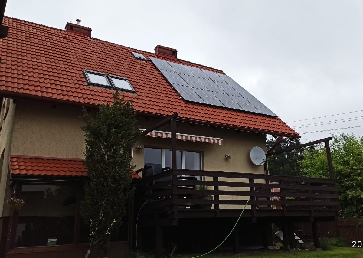 Dom jednorodzinny z czerwonym dachem, na którym zamontowane są panele słoneczne; widoczny balkon z drewnianą balustradą i markizą.