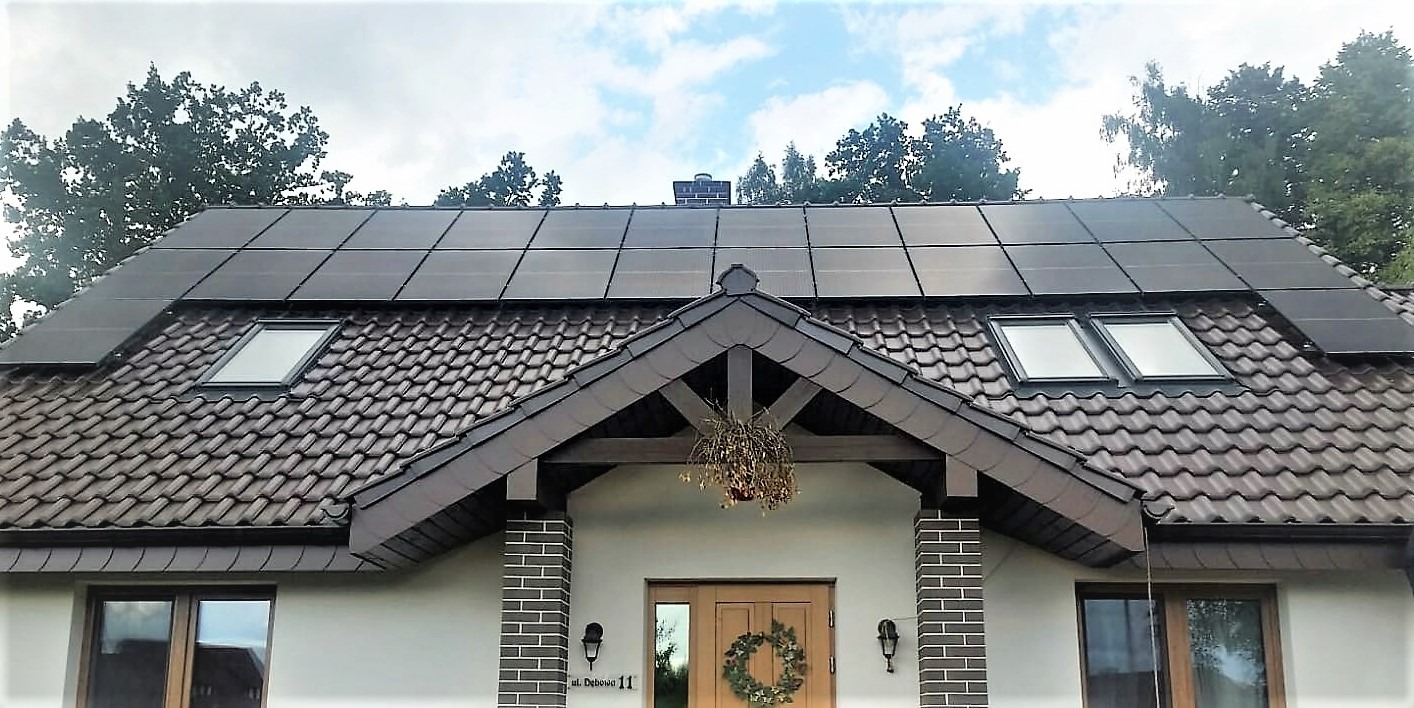 Dom jednorodzinny z ciemnymi panelami fotowoltaicznymi na brązowym dachu, widoczne okna dachowe, drewniane drzwi wejściowe z wieńcem oraz adres 'ul. Dębowa 11'.