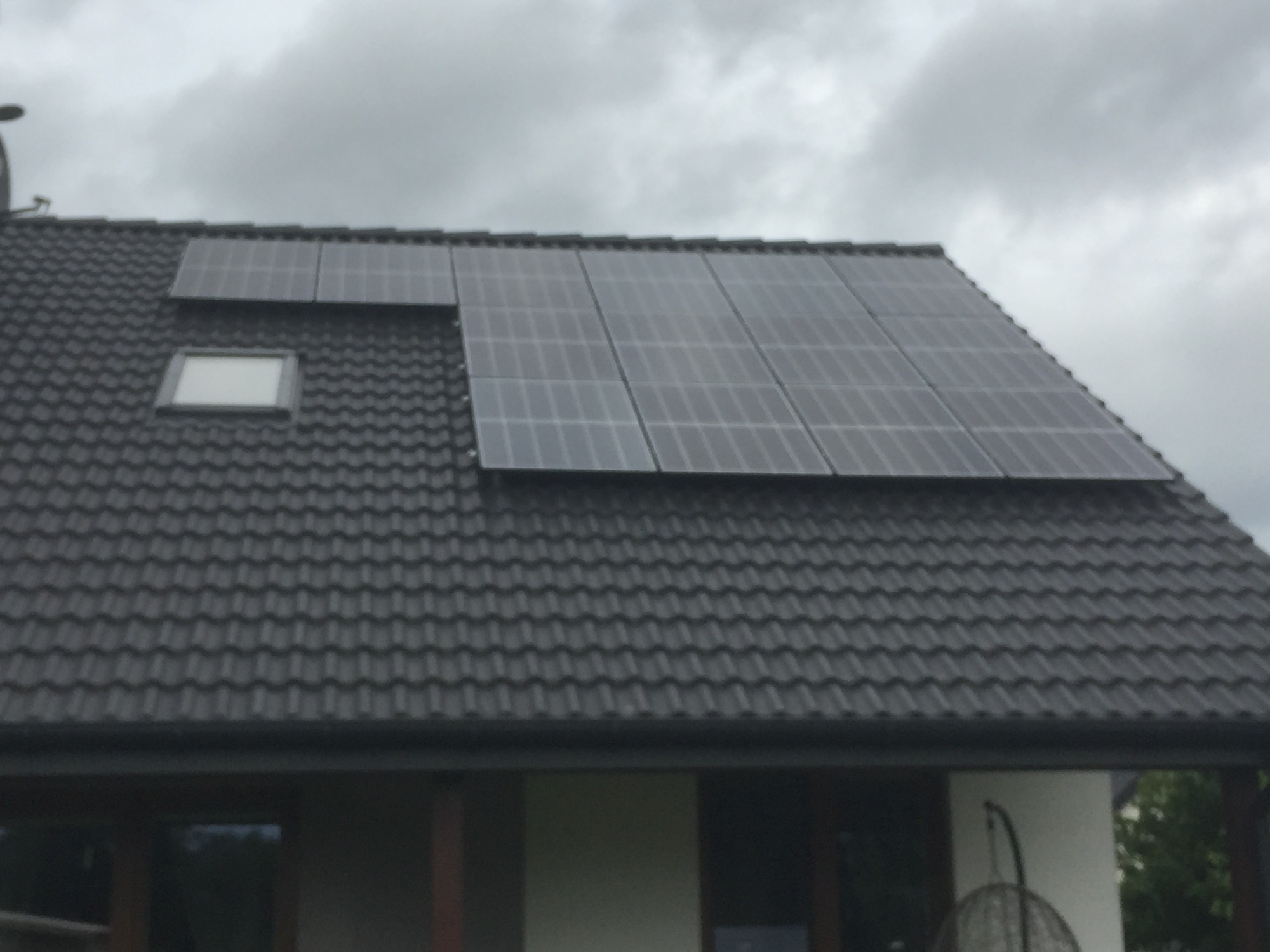 Panele słoneczne na ciemnym dachu domu jednorodzinnego z oknem dachowym, pochmurne niebo w tle.