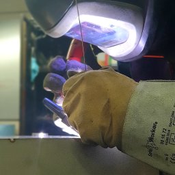 MadTom - Osoba w masce ochronnej i rękawicach spawalniczych wykonuje spoinę na metalowej powierzchni, widoczny błysk spawania i drut elektrodowy.