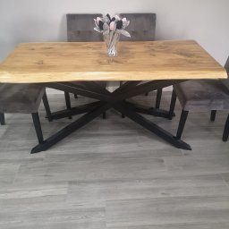 MadTom - Stół z blatem z jasnego drewna o nieregularnych krawędziach, wsparty na czarnej, metalowej podstawie o geometrycznym wzorze, otoczony szarymi krzesłami i ozdobiony wazonem z dekoracyjnymi kwiatami.