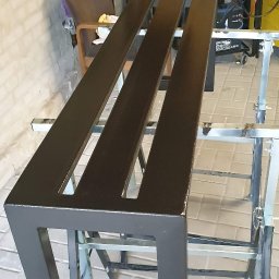 MadTom - Pomarańczowo-brązowa metalowa konstrukcja z trzema podłużnymi otworami, świeżo pomalowana, ustawiona na metalowych kozłach, w tle sprzęt spawalniczy i ściana z cegły.