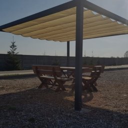 MadTom - Zadaszona pergola z rozsuwanym beżowym materiałem, pod którą znajduje się drewniany stół z ławkami, całość na żwirowym podłożu.