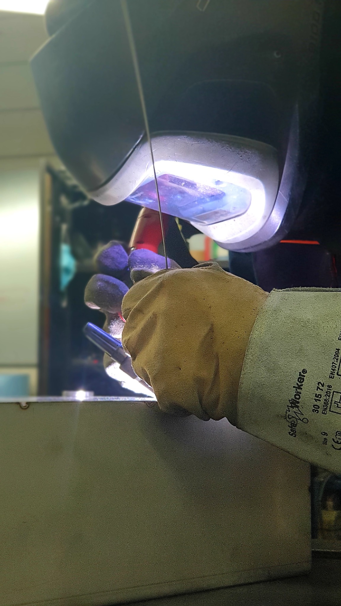 Osoba w masce ochronnej i rękawicach spawalniczych wykonuje spoinę na metalowej powierzchni, widoczny błysk spawania i drut elektrodowy.