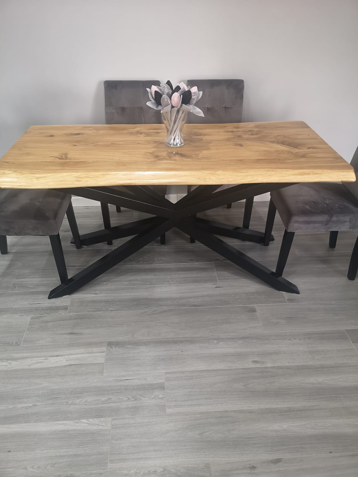 Stół z blatem z jasnego drewna o nieregularnych krawędziach, wsparty na czarnej, metalowej podstawie o geometrycznym wzorze, otoczony szarymi krzesłami i ozdobiony wazonem z dekoracyjnymi kwiatami.