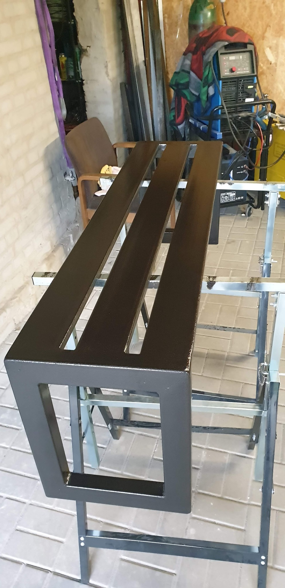 Pomarańczowo-brązowa metalowa konstrukcja z trzema podłużnymi otworami, świeżo pomalowana, ustawiona na metalowych kozłach, w tle sprzęt spawalniczy i ściana z cegły.