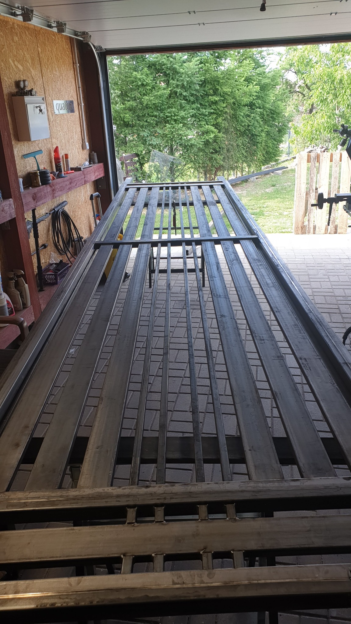 Metalowa konstrukcja bramy w trakcie tworzenia, widoczne spawy i elementy konstrukcyjne, tło warsztatowe z otwartą bramą garażową i widokiem na zieleń.