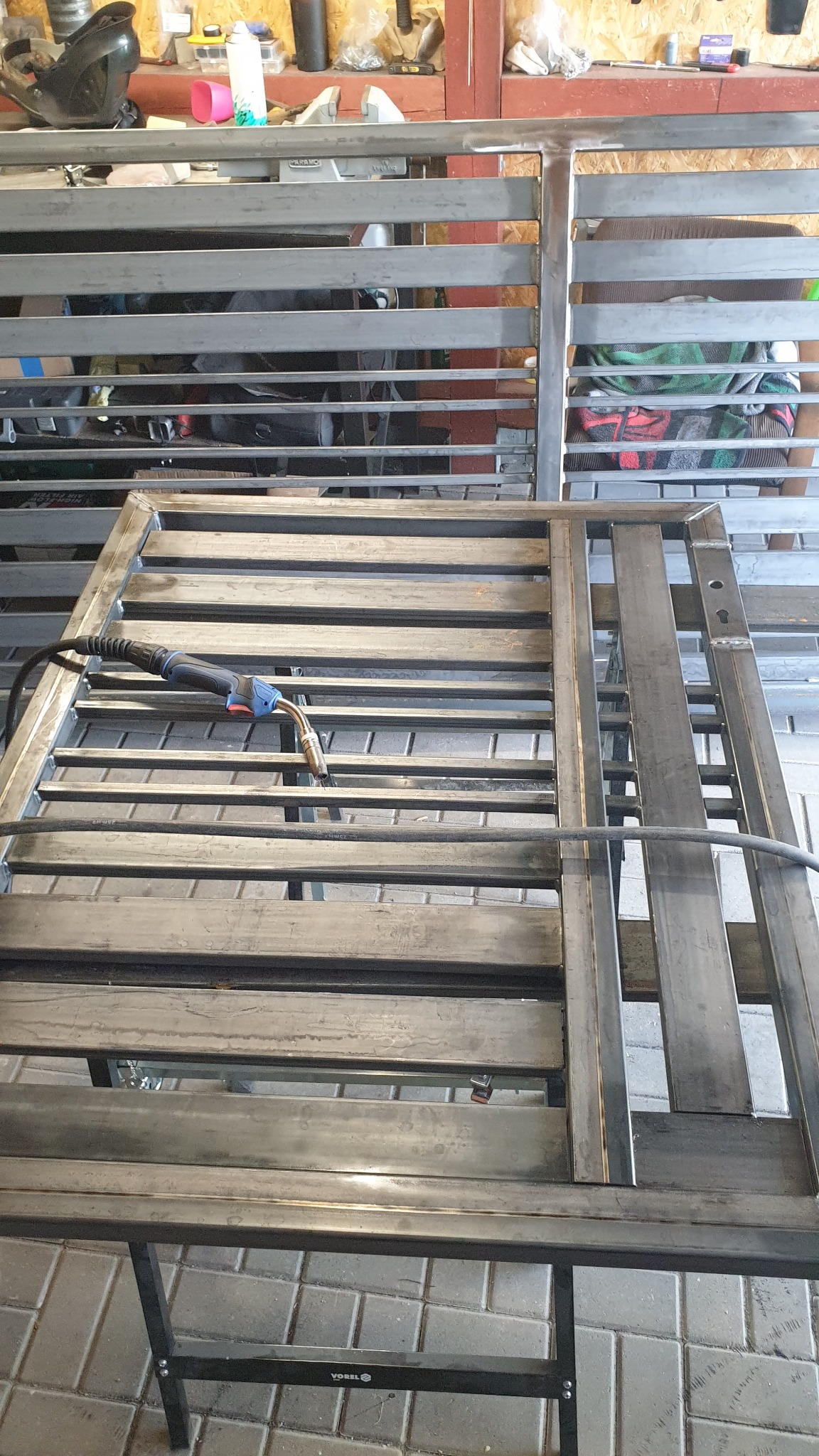 Spawanie metalowej konstrukcji z poziomymi elementami, widoczny uchwyt spawarki Mig/Mag, konstrukcja umieszczona na stole roboczym.