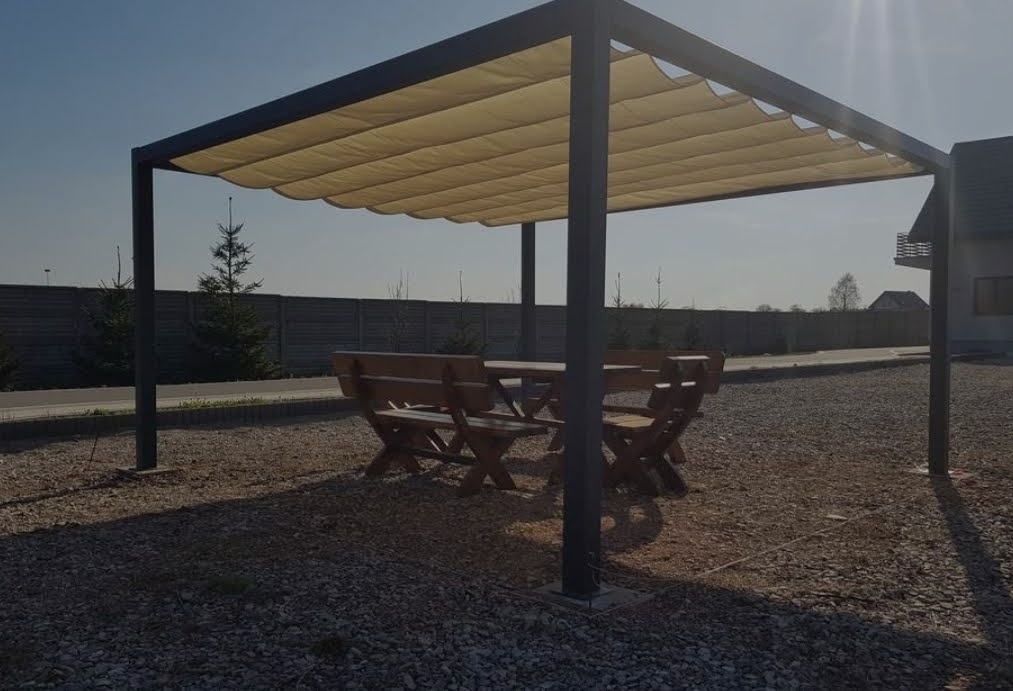 Zadaszona pergola z rozsuwanym beżowym materiałem, pod którą znajduje się drewniany stół z ławkami, całość na żwirowym podłożu.