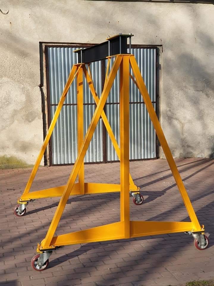 Żółty, mobilny portalowy dźwig warsztatowy na kółkach, z czarną belką poprzeczną, na tle metalowych drzwi i ściany budynku.