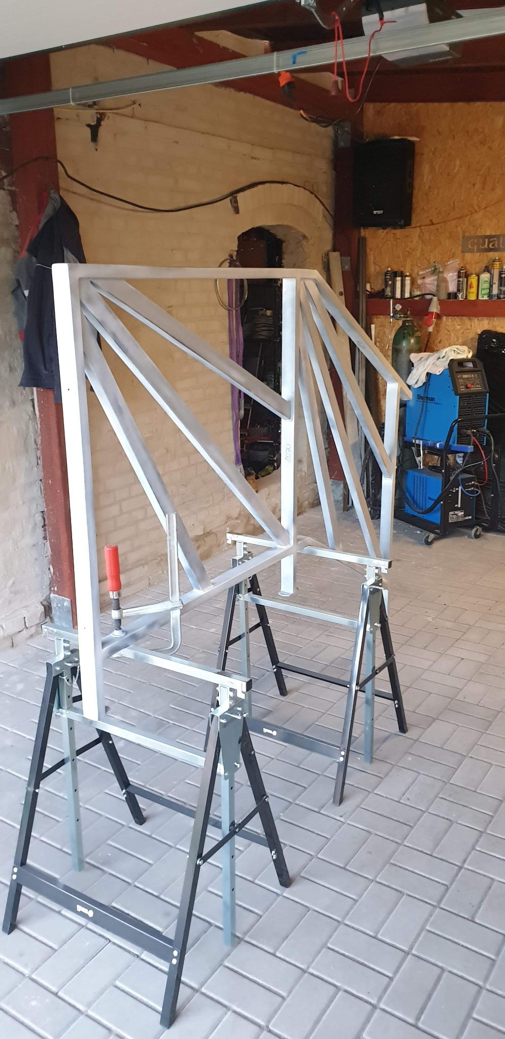 Metalowa konstrukcja schodów z poręczą, wstępnie pomalowana na szaro, zamocowana na dwóch kozłach warsztatowych w garażu. W tle widoczny sprzęt spawalniczy i inne narzędzia.