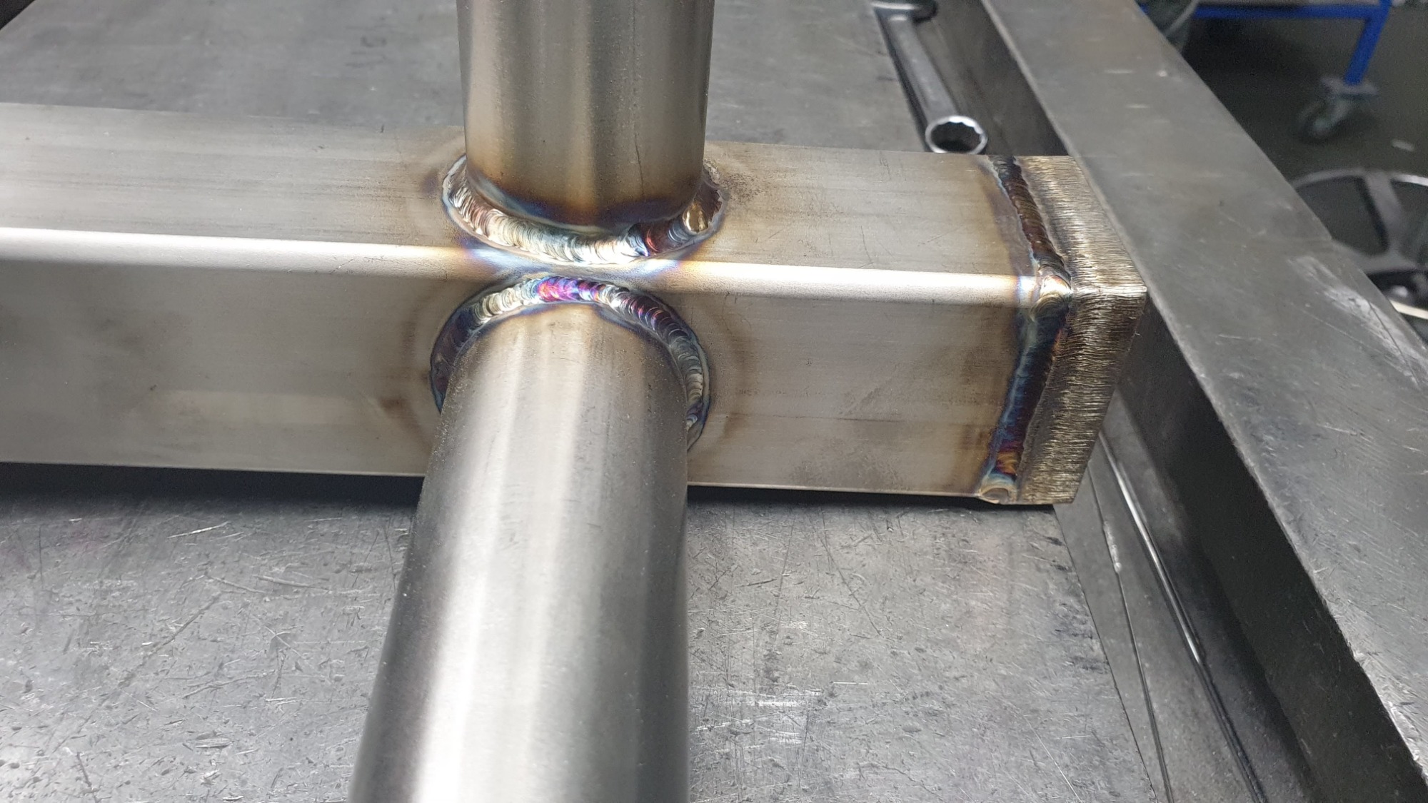 Spoina łącząca dwie rury ze stalowym profilem kwadratowym, widoczne przebarwienia termiczne na spoinie i powierzchni metalu, klucz płaski w tle.